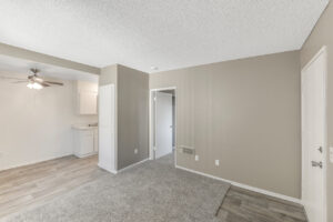 Interior Unit Living Room, neutral toned wall paint and carpeting, bedroom entryway adjacent to apartment entrance.